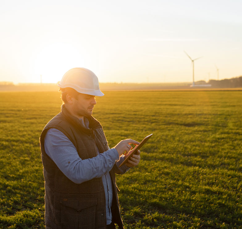 Engenheiro realiza a avaliação de imóveis rurais usando um tablet em um campo com turbinas de energia eólica ao fundo.