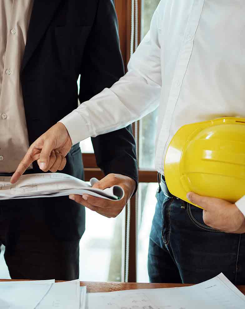 dois homens em pé, um de terno escuro e outro de camisa branca segurando um capacete amarelo, apontam para plantas arquitetônicas sobre uma mesa, ilustrando a análise conjunta na validação de um laudo de avaliação de imóvel para desapropriação